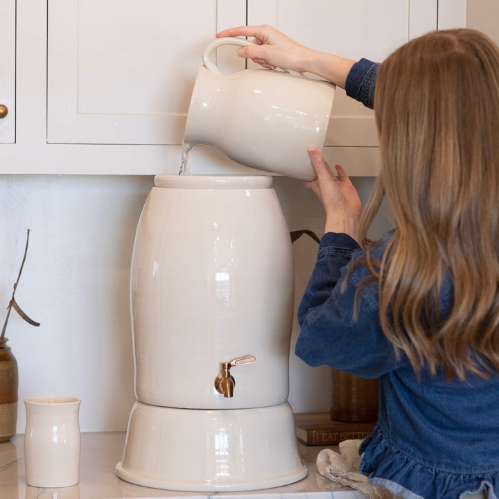 Person pouring water from a white pitcher into a white ceramic water filter.