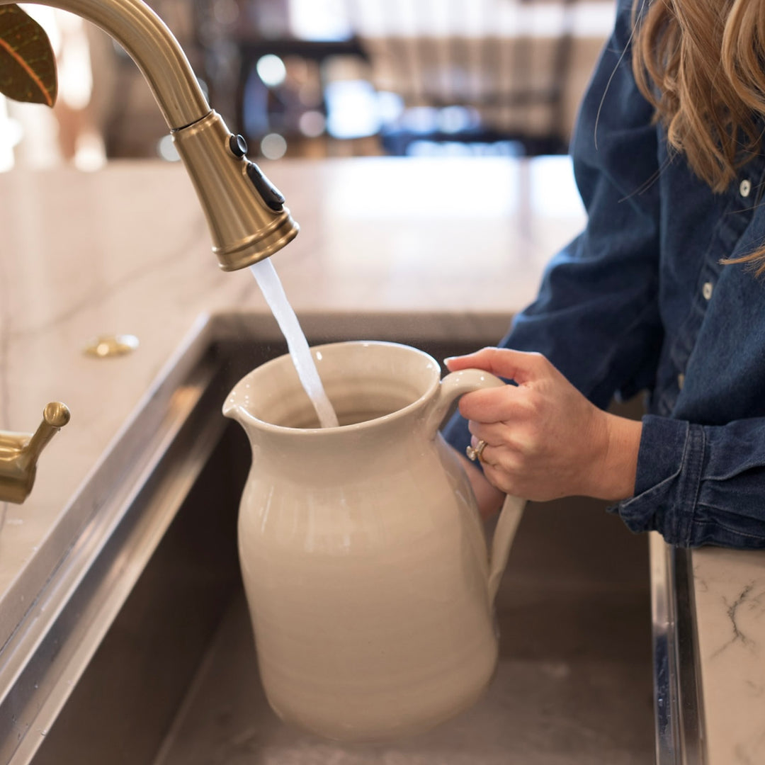 Person filling a white ceramic pitcher with water from a brass faucet in a kitchen.