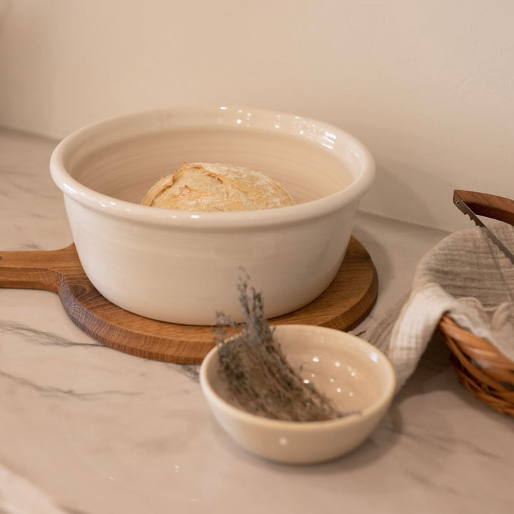 White ceramic bowl storing bread in a kitchen with a bowl of fresh herbs, and a woven basket on a marble surface.