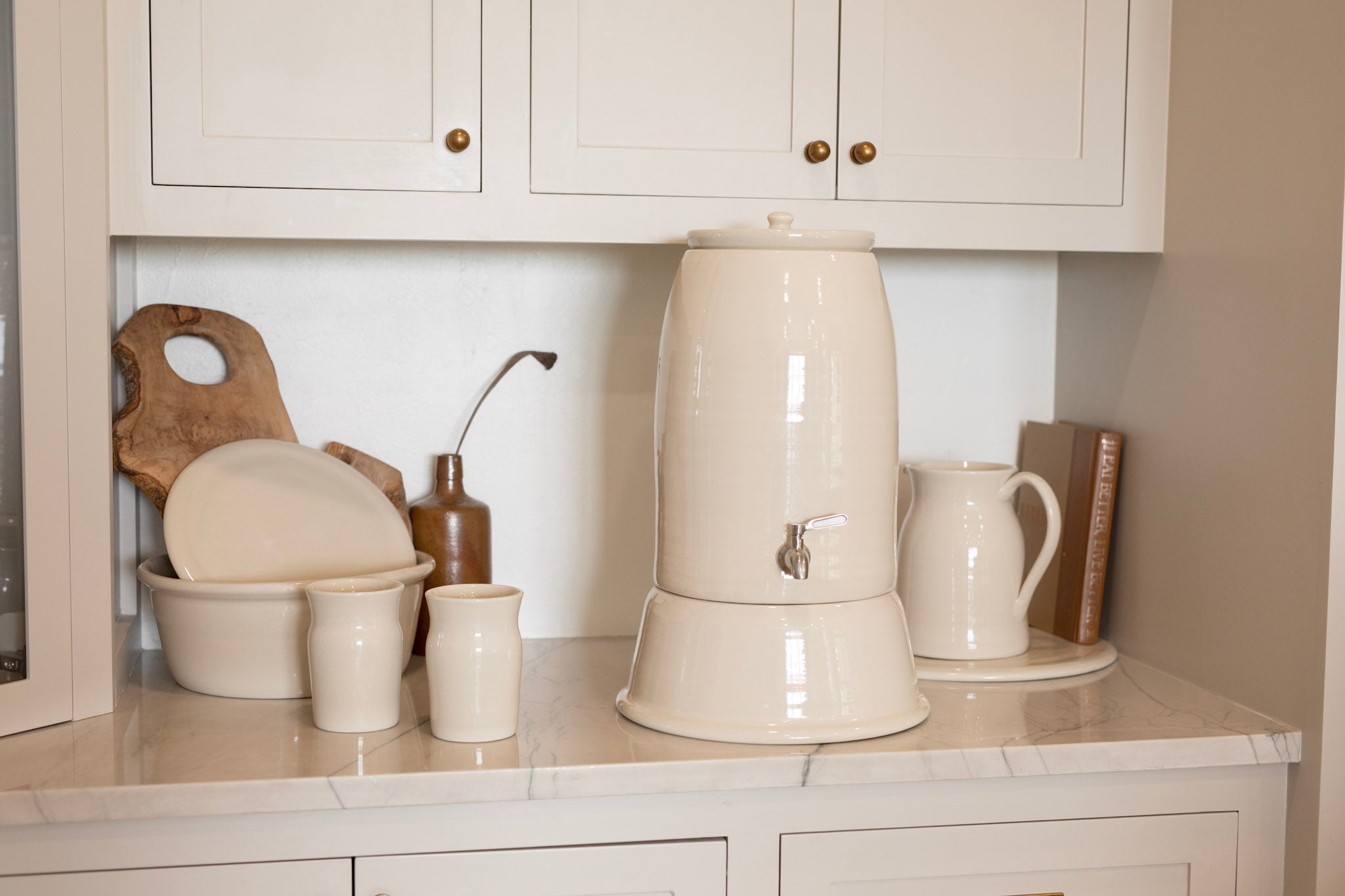 A cream-colored ceramic water dispenser with a built-in lid and metal spout, displayed on a marble-look countertop alongside matching ceramic cups, a white pitcher, and wooden kitchen accessories under white cabinets.