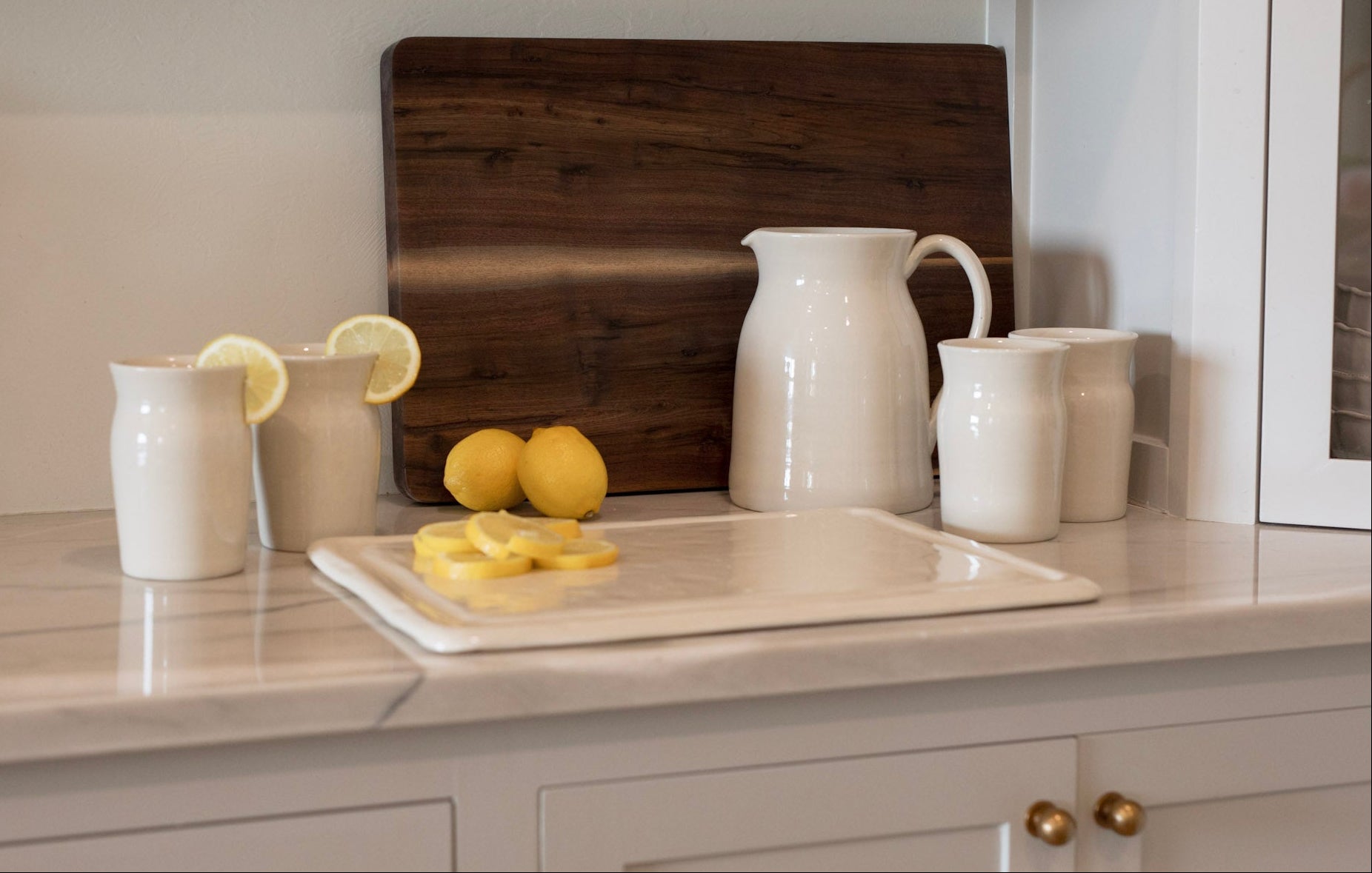 A set of white ceramic kitchenware, including pitchers, mugs, and a serving tray, arranged on a marble countertop with lemons and a wooden cutting board in the background.