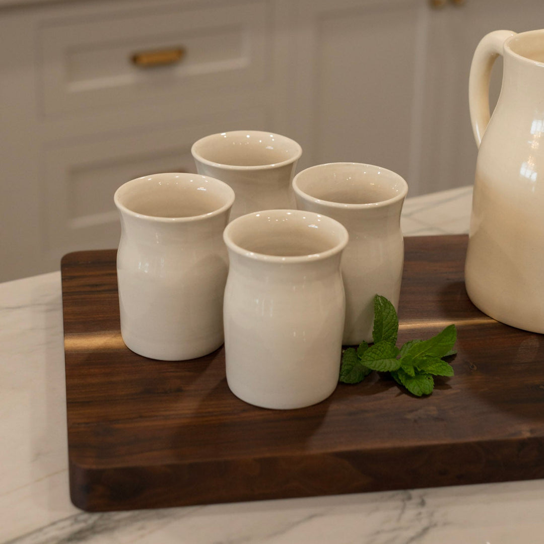 Set of four ceramic cups on a wooden cutting board with a pitcher in a kitchen setting.