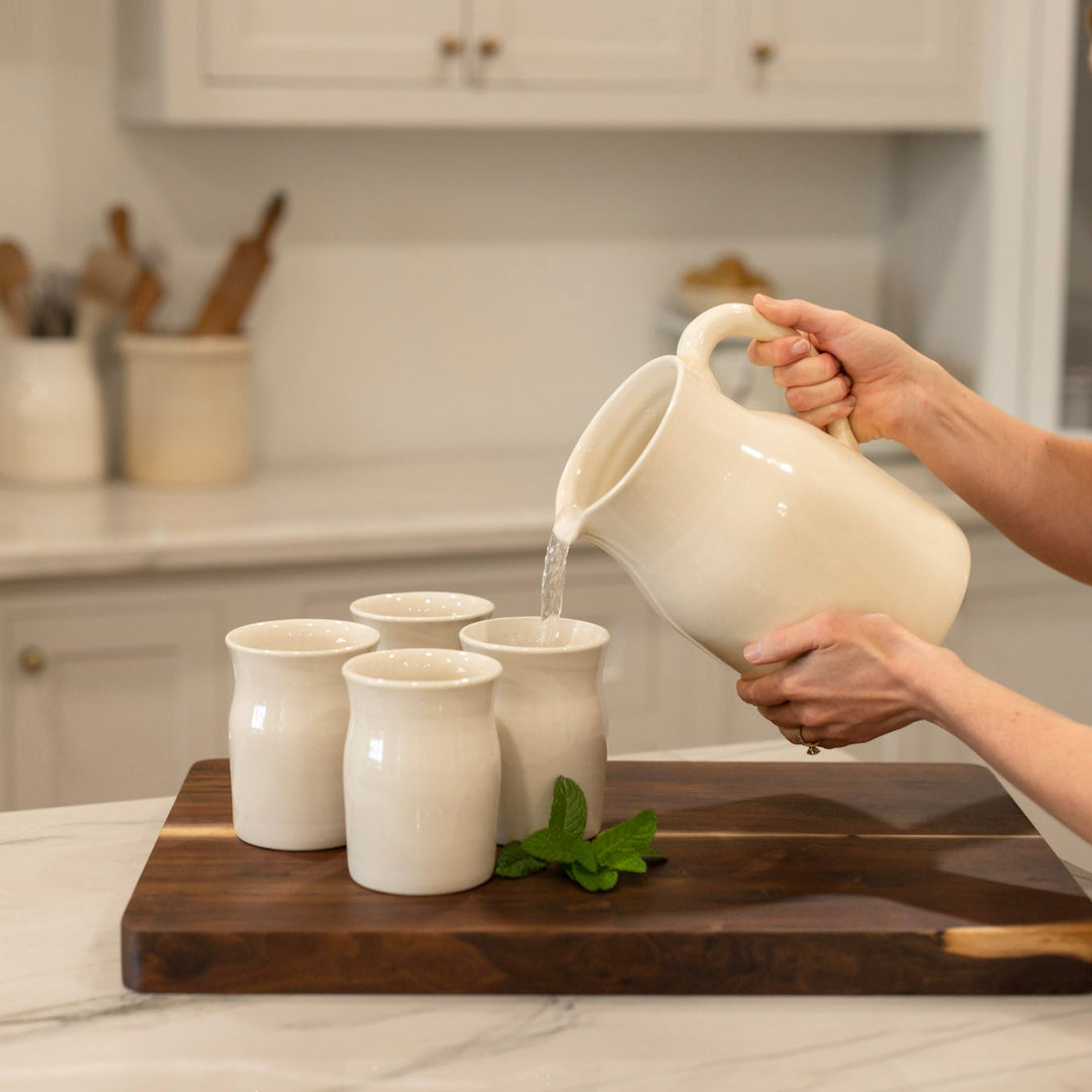 Person pouring water from a ceramic pitcher into ceramic cups on a wooden cutting board in a kitchen.