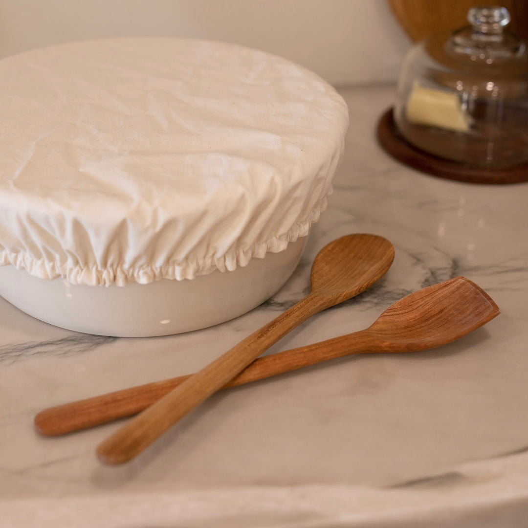Two hand scraped wooden spoons on a marble surface with a covered dish in the background.