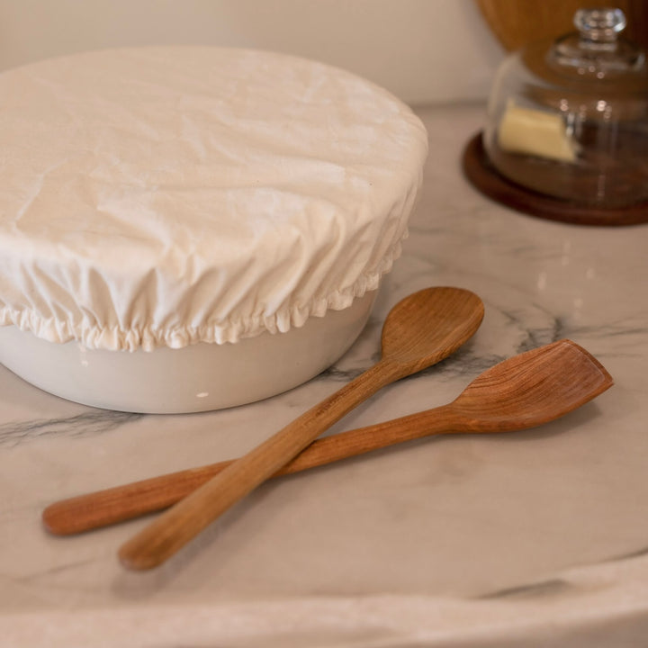 Two hand scraped wooden spoons on a marble surface with a covered dish in the background.