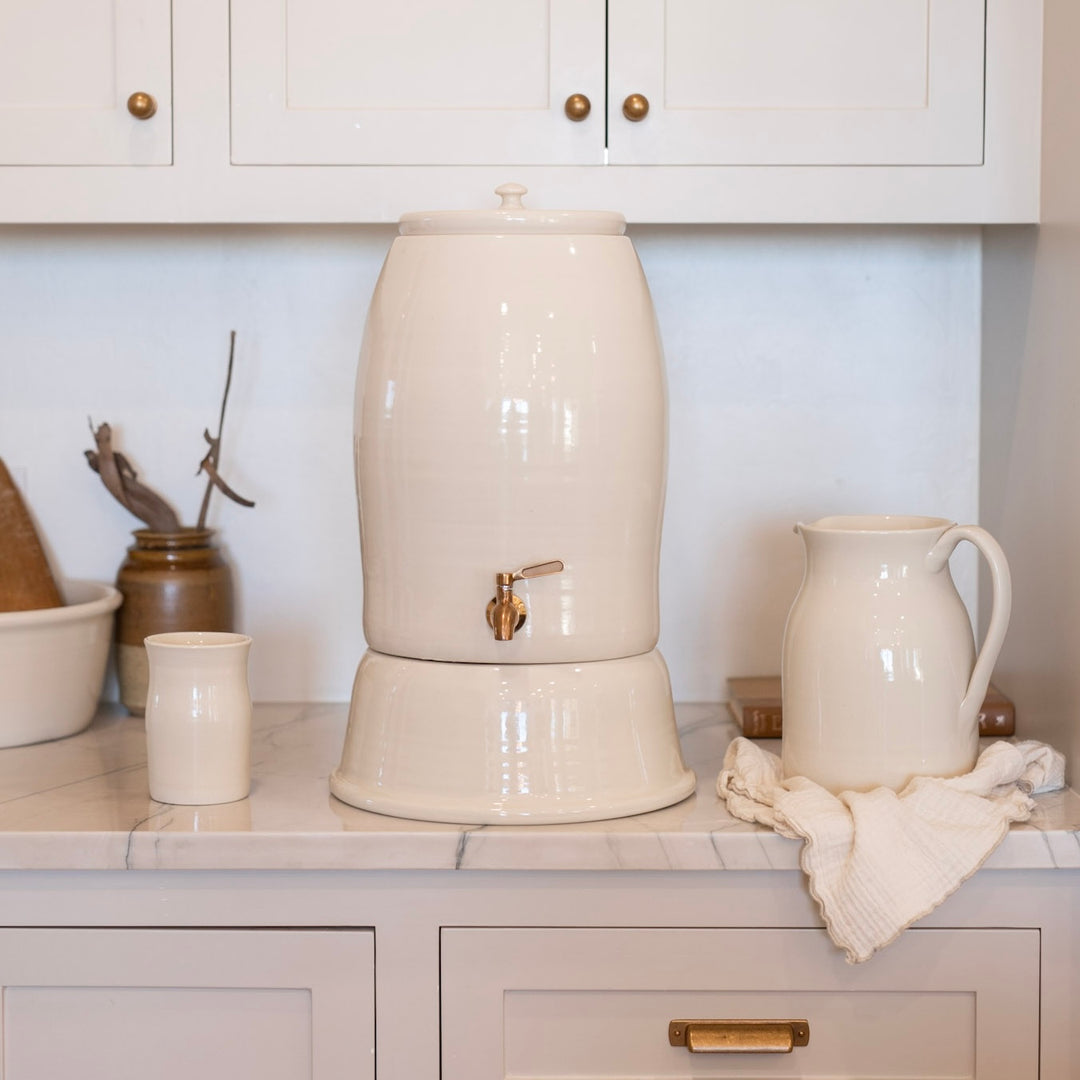 White ceramic water dispenser on a kitchen counter with a water pitcher, water cups, and a towel.