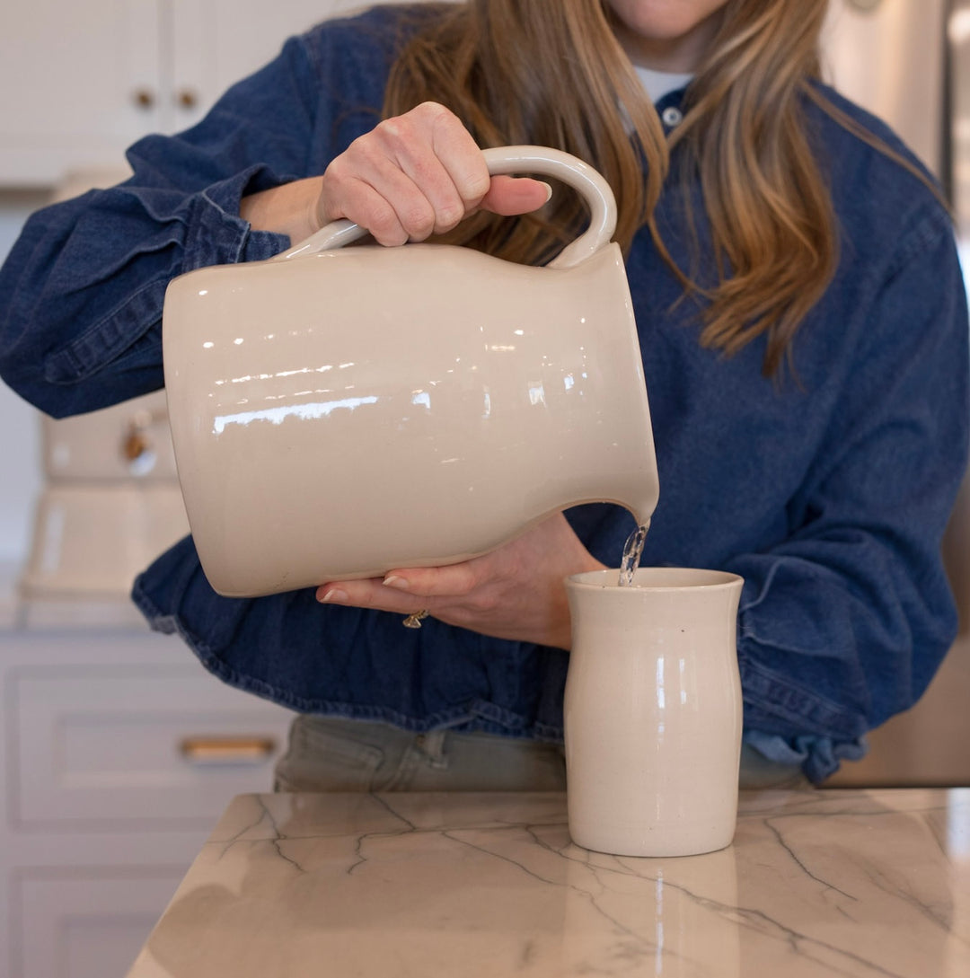 Person pouring water from a large white pitcher into a ceramic cup on a marble countertop.